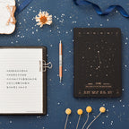 Flat lay of a black hardcover Aries zodiac notebook with a gold star map constellation design, an open journal with handwritten Chinese text, and a mechanical pen on a blue starry desk mat.
