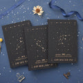 Three black hardcover notebooks featuring gold star constellation designs for Leo, Scorpio, and Aquarius zodiac signs on a blue background.
