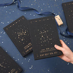 A person holding a black hardcover Gemini zodiac notebook with a gold star constellation design alongside a matching Sagittarius notebook on a blue starry surface.