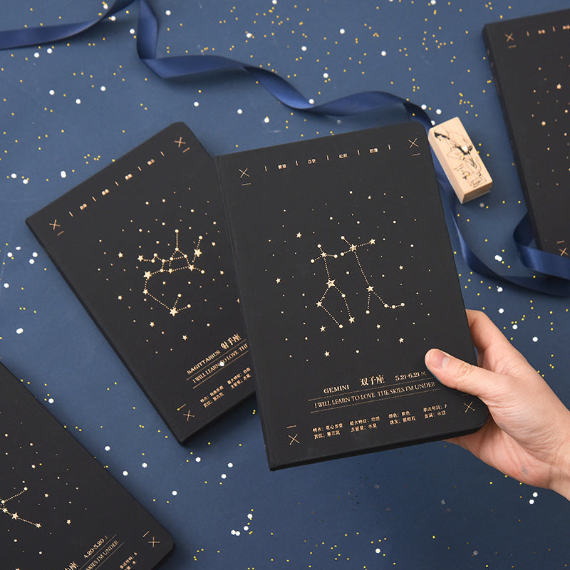 A person holding a black hardcover Gemini zodiac notebook with a gold star constellation design alongside a matching Sagittarius notebook on a blue starry surface.