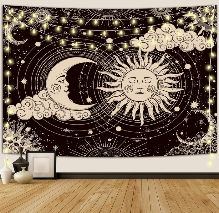 Aesthetic black and white celestial tapestry featuring a mystic sun and a crescent moon with faces, surrounded by stars, clouds, and hanging fairy lights on a bedroom wall.