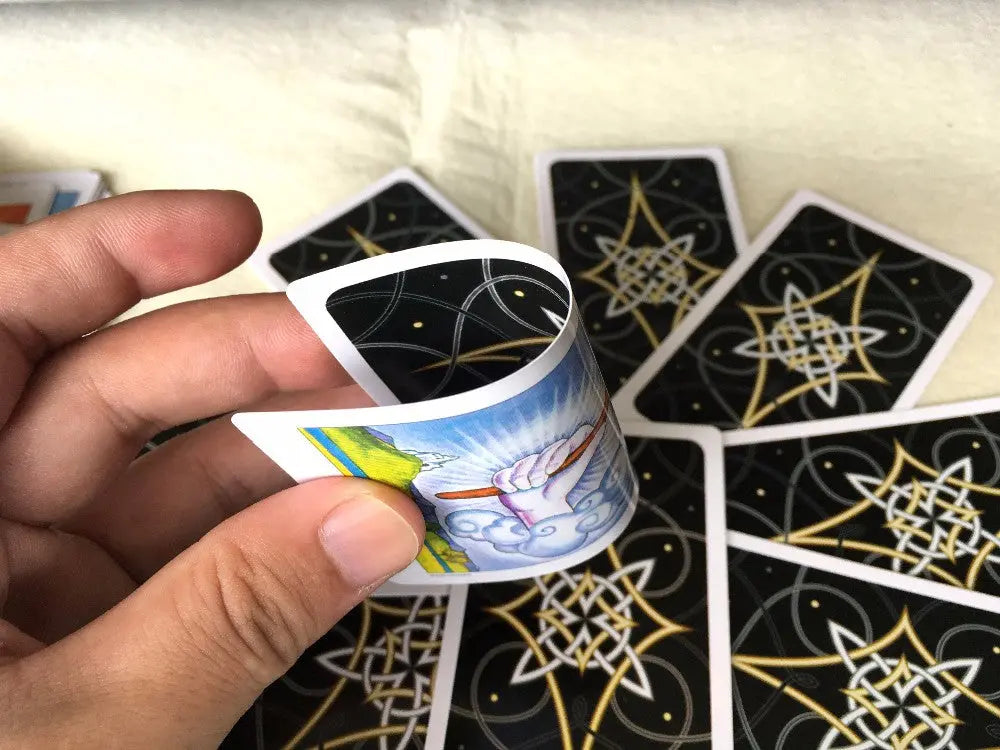 A close-up view of a hand squeezing and bending a tarot card completely in half to demonstrate its flexible paper elasticity. The underside reveals a classic Rider-Waite-Smith Ace of Wands illustration, while the background shows a spread of black card backs decorated with a golden geometric star design.
