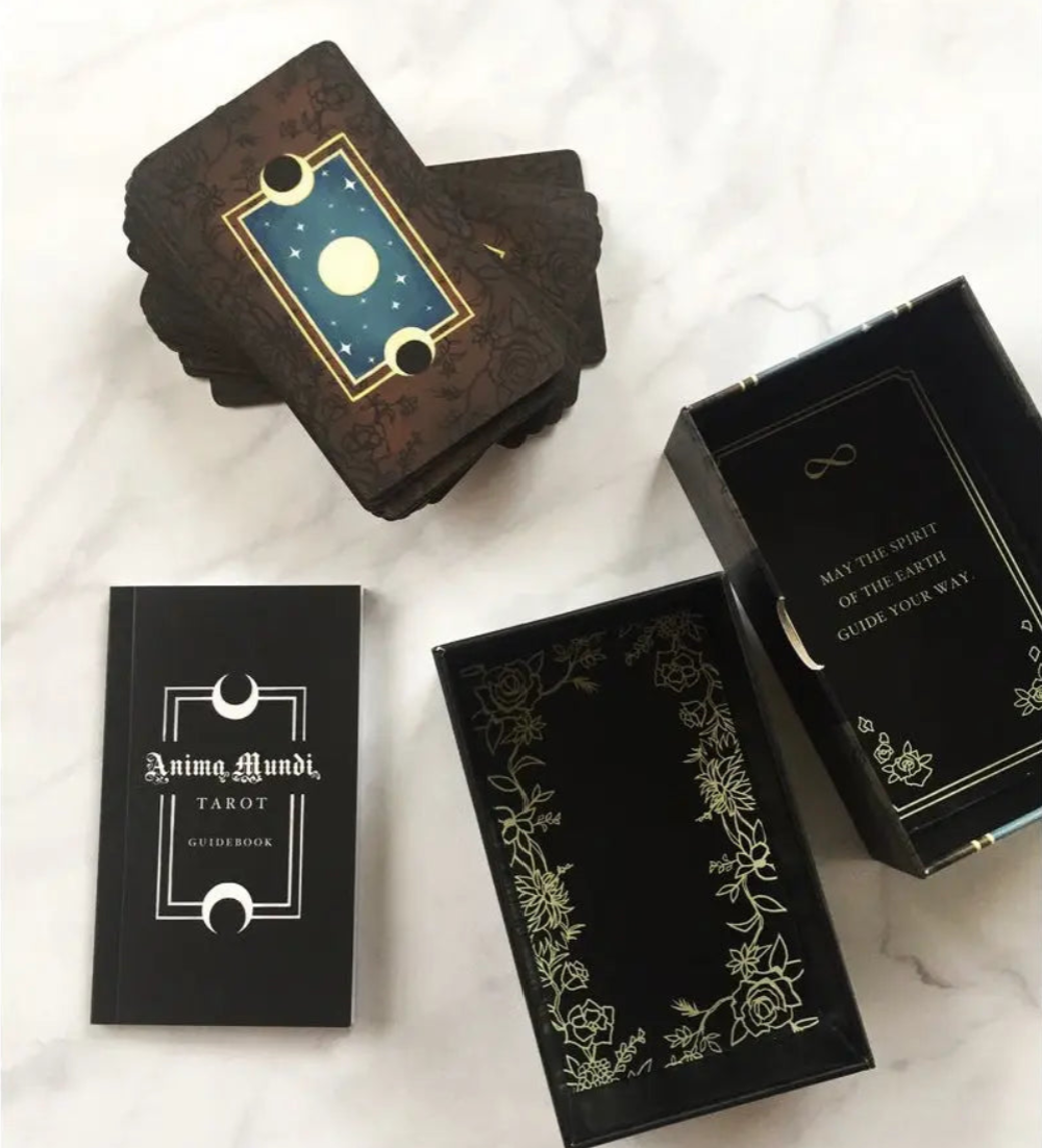 An open two-piece rigid Anima Mundi box next to gold-edged cards and a physical booklet guide.