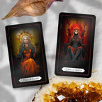 Two tarot cards resting diagonally on a light surface accented by a dried leaf and a golden crystal cluster. The left card shows the Queen of Coins holding a mechanical spiked sphere beneath a bright sun halo, while the right card displays the skull-masked Queen of Wands seated on a fiery throne.