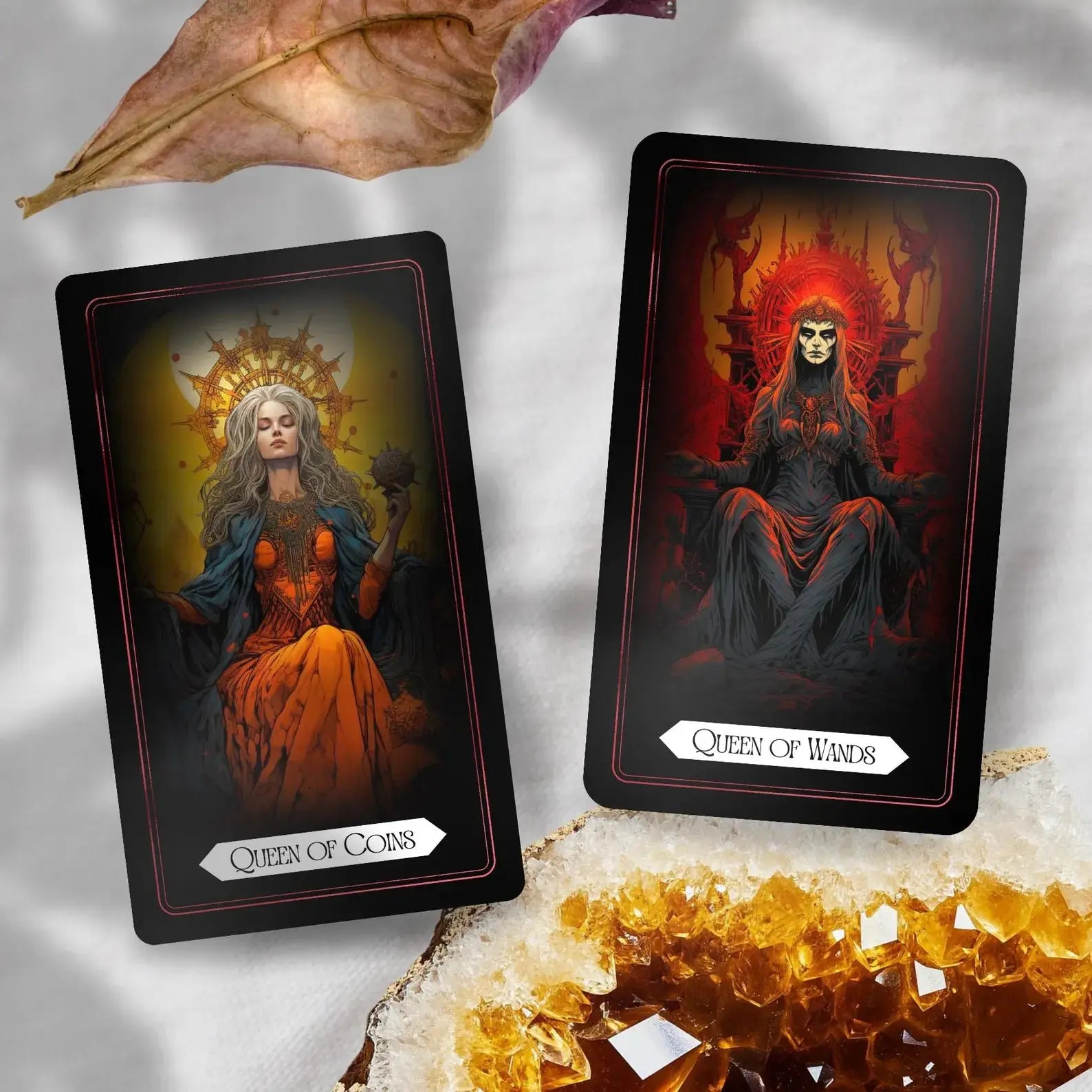 Two tarot cards resting diagonally on a light surface accented by a dried leaf and a golden crystal cluster. The left card shows the Queen of Coins holding a mechanical spiked sphere beneath a bright sun halo, while the right card displays the skull-masked Queen of Wands seated on a fiery throne.