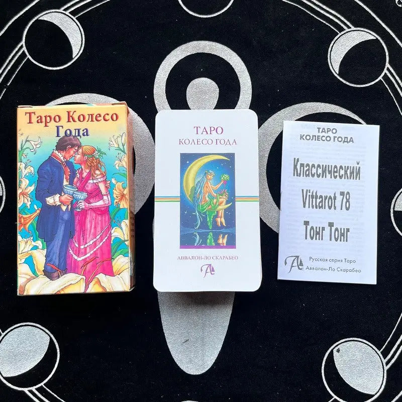A colorful card box labeled "Таро Колесо Года" displaying a romantic couple holding chalices. To its right sits a card depicting a celestial figure on a crescent moon, and a white booklet titled "Классический Vittarot 78 Тонг Тонг", all resting on a black patterned altar cloth.