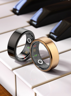 A shiny black smart ring and a gold smart ring balancing on white piano keys, showing inner sensor arrays.