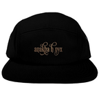 Black five-panel camper style hat featuring the elegant Anakha H Nyx calligraphy script logo embroidered in a warm copper bronze tone across the front.