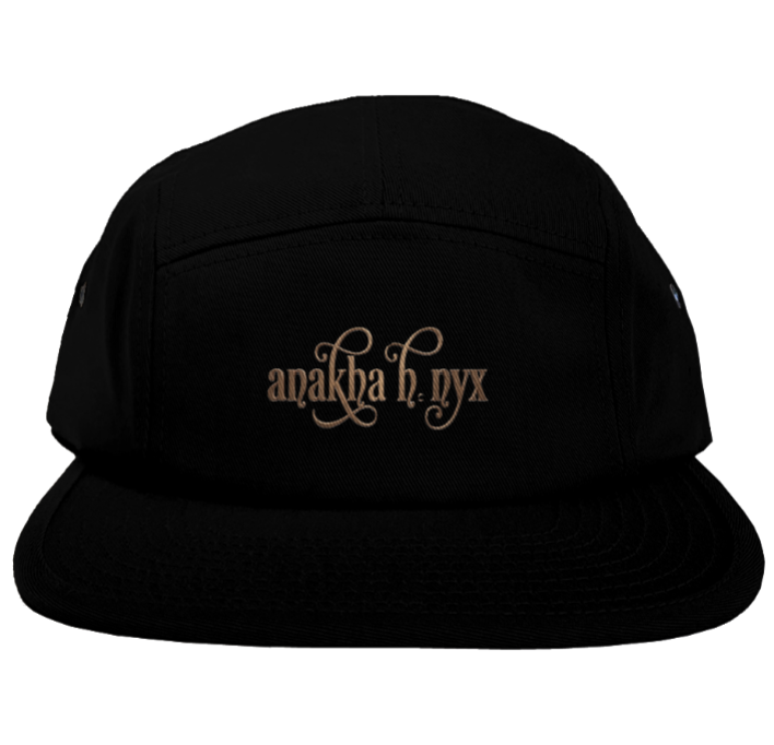 Black five-panel camper style hat featuring the elegant Anakha H Nyx calligraphy script logo embroidered in a warm copper bronze tone across the front.