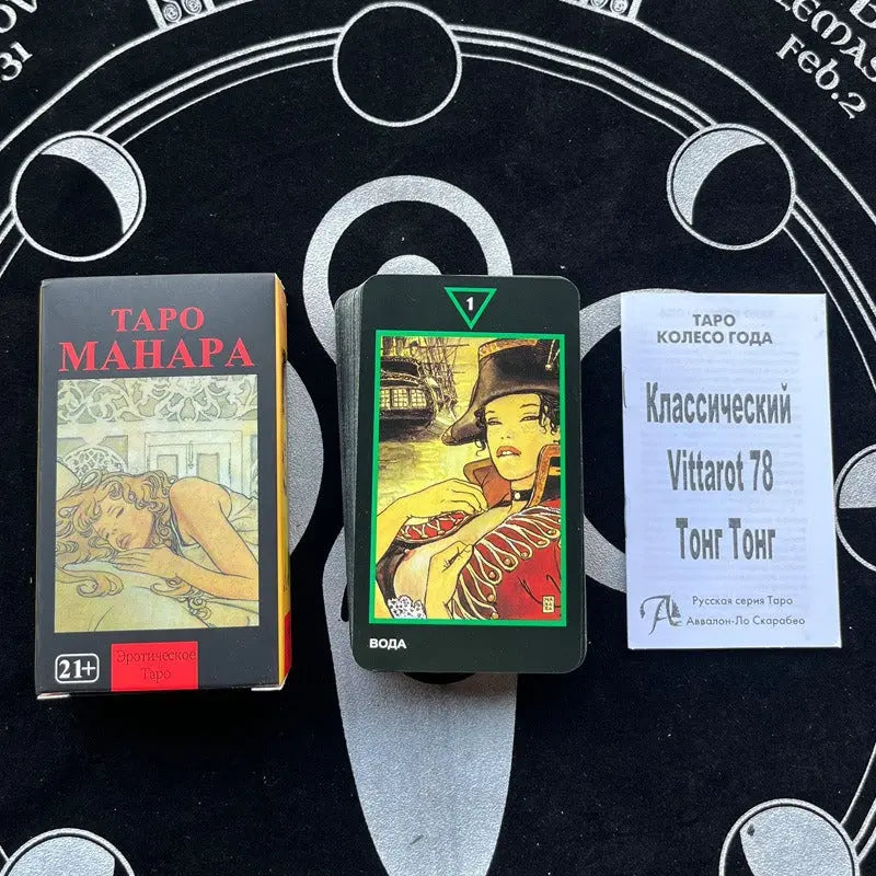 Russian Language Manara Erotic A dark grey card box labeled "ТАРО МАНАРА" featuring a "21+" age restriction badge and a graphic illustration of a reclining woman. To its right rests a card displaying a woman in military attire with a green triangular elemental crest labeled "ВОДА" (Water), next to a white booklet titled "Классический Vittarot 78 Тонг Тонг" on a dark cloth.