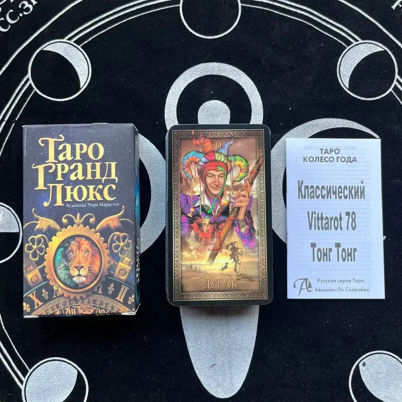 A Tarot Grand Luxe box with a lion illustration ("Таро Гранд Люкс") sits next to a card showing a colorful jester labeled "ДУРАК" (The Fool). A small white paper insert titled "Классический Vittarot 78 Тонг Тонг" rests beside them on a black patterned surface.