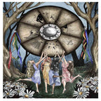 An full-page watercolor and ink artistic illustration from a book depicting a seasonal pagan ritual. In a dark, twilight forest setting flanked by large barren trees, four women with long hair dance in an active circle with their arms raised toward a massive wooden wheel floating above them. The women wear flowing dresses in light blue, golden yellow, purple, and pale pink, and white daisies are scattered across the grassy ground in the foreground. Hanging from the trees on both sides of the floating wheel 