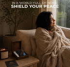 A cozy lifestyle marketing photograph showcasing a woman relaxing on a couch wrapped in a chunky knit blanket next to a wooden side table with the Anima Protection Crystals set. The tabletop setup features the open wooden crystal box, a black glass candle jar, and a branded guide booklet under the overlay text Shield Your Peace.