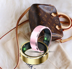 Pink and gold smart health rings with internal circuitry showing, placed in front of a luxury designer handbag.