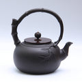 This authentic Japanese Tetsubin cast iron teapot features a structural circular bamboo-joint overhead handle paired with a matching bamboo-spout design. The heavy-duty stovetop tea kettle displays an embossed raised bamboo leaf motif on its matte black body and a flat lid with a decorative pumpkin-shaped top knob.