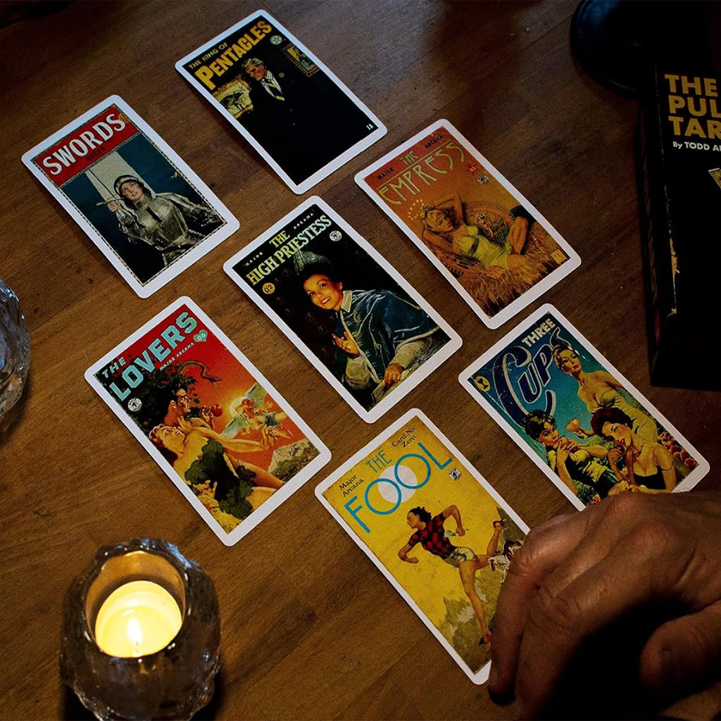 This lifestyle presentation of The Pulp Tarot showcases a dynamic multi-card reading laid out on a rustic wooden table illuminated by candlelight. The atmospheric view features high-detail mid-century illustration designs across seven archetypes including the King of Pentacles, Swords, The Empress, The High Priestess, The Lovers, Three of Cups, and The Fool.