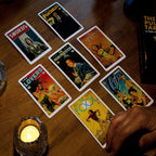 This lifestyle presentation of The Pulp Tarot showcases a dynamic multi-card reading laid out on a rustic wooden table illuminated by candlelight. The atmospheric view features high-detail mid-century illustration designs across seven archetypes including the King of Pentacles, Swords, The Empress, The High Priestess, The Lovers, Three of Cups, and The Fool.