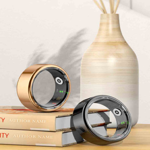A lifestyle product shot showcasing two health tracker smart rings in polished rose gold and glossy black finishes. The rings rest on stacked books next to a ceramic vase, displaying their inner resin lining packed with visible electronic sensors and bio-tracking health components.