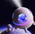 A lifestyle product rendering of the TOPINCN mini spaceship desk humidifier emitting a fine ultrasonic cool mist spray into a dark outer space setting. The purple capsule body glows with a colorful ambient LED light ring while a matching miniature astronaut figurine stands on a lunar planetary surface below.