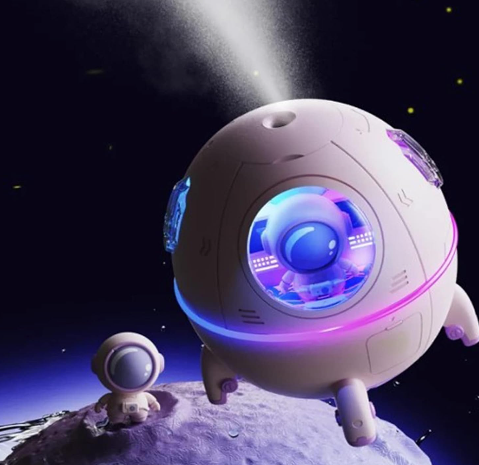A lifestyle product rendering of the TOPINCN mini spaceship desk humidifier emitting a fine ultrasonic cool mist spray into a dark outer space setting. The purple capsule body glows with a colorful ambient LED light ring while a matching miniature astronaut figurine stands on a lunar planetary surface below.
