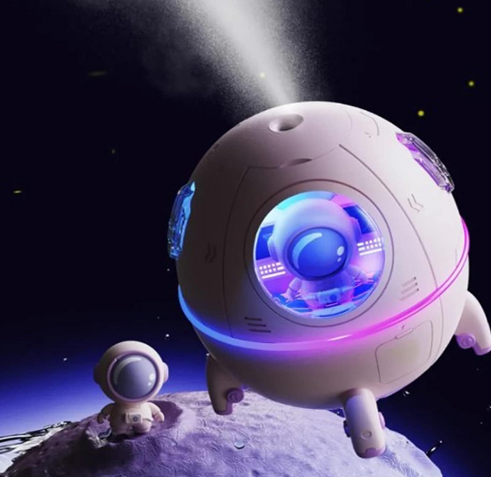 A lifestyle product rendering of the TOPINCN mini spaceship desk humidifier emitting a fine ultrasonic cool mist spray into a dark outer space setting. The purple capsule body glows with a colorful ambient LED light ring while a matching miniature astronaut figurine stands on a lunar planetary surface below.
