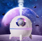 A lifestyle graphic of the TOPINCN spaceship ultrasonic cool mist humidifier emitting a thick plume of water vapor from its top spout against a cosmic space background. The purple capsule features an integrated glowing LED ring light and a clear front visor revealing the small astronaut figurine inside